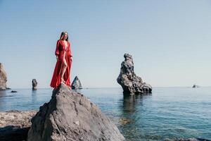 Frau Reise Meer. jung glücklich Frau im ein lange rot Kleid posieren auf ein Strand in der Nähe von das Meer auf Hintergrund von vulkanisch Felsen, mögen im Island, Teilen Reise Abenteuer Reise foto