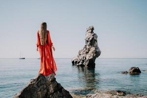 Frau Reise Meer. jung glücklich Frau im ein lange rot Kleid posieren auf ein Strand in der Nähe von das Meer auf Hintergrund von vulkanisch Felsen, mögen im Island, Teilen Reise Abenteuer Reise foto