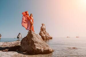Frau Reise Meer. jung glücklich Frau im ein lange rot Kleid posieren auf ein Strand in der Nähe von das Meer auf Hintergrund von vulkanisch Felsen, mögen im Island, Teilen Reise Abenteuer Reise foto