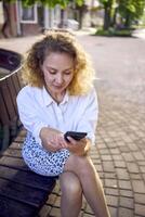 ein schön Mitte Alter Frau im 70er, 80er Jahre Stil Kleider ist suchen beim ihr Smartphone auf ein Bank foto