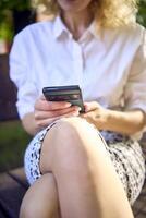 ein schön Mitte Alter Frau im 70er, 80er Jahre Stil Kleider ist suchen beim ihr Smartphone auf ein Bank foto