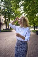 ein schön Mitte Alter Frau im 70er, 80er Jahre Stil Kleider erstreckt sich nach Arbeit auf ein Sonne durchnässt Gasse foto