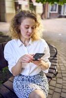 ein schön Mitte Alter Frau im 70er, 80er Jahre Stil Kleider ist suchen beim ihr Smartphone auf ein Bank foto