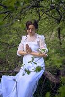 ein schön Frau im ein Weiß Jahrgang Kleid mit ein Zug ist beten während Sitzung auf ein gefallen Baum foto