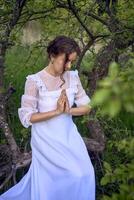 ein schön Frau im ein Weiß Jahrgang Kleid mit ein Zug ist beten während Sitzung auf ein gefallen Baum foto