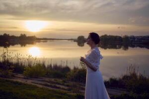 ein Frau im ein Weiß Jahrgang Kleid auf das Ufer von ein See beim Sonnenuntergang foto