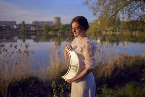 ein Frau im ein Weiß Jahrgang Kleid auf das Ufer von ein See beim Sonnenuntergang hält ein Mond geformt Spiegel foto