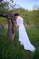 ein schön Frau im ein Weiß Jahrgang Kleid mit ein Zug ist streicheln ein Baum beschädigt durch ein Sturm foto