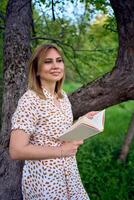 Frau im ein Pastell- Kleid liest ein Buch in der Nähe von ein Baum im das Garten foto