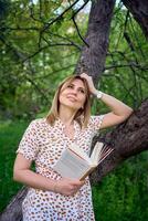 Frau im ein Pastell- Kleid liest ein Buch in der Nähe von ein Baum im das Garten foto