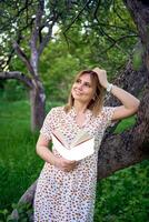 Frau im ein Pastell- Kleid liest ein Buch in der Nähe von ein Baum im das Garten, Attrappe, Lehrmodell, Simulation foto