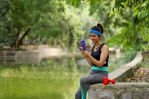 passen weiblich Sitzung im Park halten Flasche im einer Hand und Telefon im andere Hand foto