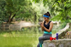 überrascht Jogger Dame Sitzung Fluss neben im Fitnessstudio passen suchen beim Handy foto