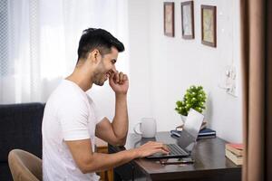 gut aussehend jung Kerl im Weiß Outfit Sitzung auf Stuhl Herstellung Anruf auf seine Laptop foto