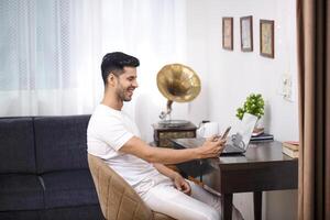 glücklich lächelnd Mann im Weiß Outfit lesen Botschaft auf Telefon während Arbeiten beim Zuhause Büro foto