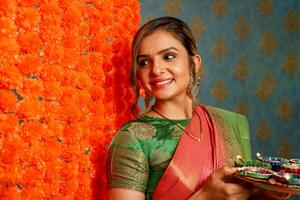 ein schüchtern Mädchen im Saree halten Diya Teller während Stehen im Vorderseite von ein Blumengeschmückt Mauer foto