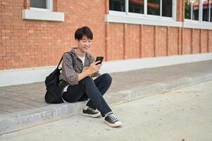 ein positiv asiatisch männlich Hochschule Schüler sitzt auf das Straße in der Nähe von das Campus Gebäude mit seine Telefon. foto