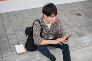 ein positiv asiatisch männlich Hochschule Schüler sitzt auf das Straße in der Nähe von das Campus Gebäude mit seine Telefon. foto