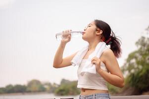 ein jung asiatisch Frau Getränke von ein Wasser Flasche, Kühlung Nieder nach Gehen draußen im ein heiß Tag. foto