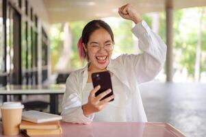 ein aufgeregt asiatisch weiblich Hochschule Schüler feiert gut Nachrichten während suchen beim ihr Telefon beim ein Tisch. foto