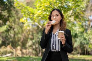 ein Geschäftsfrau ist haben ein schnell Frühstück im ein Stadt Park, Essen ein Sandwich und Trinken Kaffee. foto