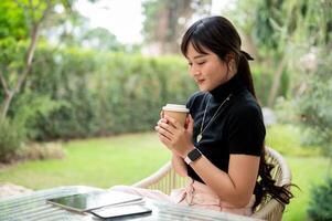 ein Schön, positiv asiatisch Frau sitzt beim ein draussen Tabelle im ein Grün Garten genießen Kaffee. foto