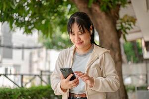 jung asiatisch Frau tragen Ohrhörer, mit ihr Telefon, Hören zu Musik- während chillen im ein Stadt Park foto