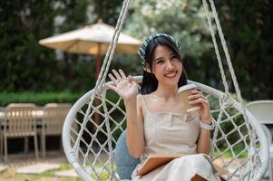 ein attraktiv asiatisch Frau chillen auf ein schwingen im ein Garten, winken ihr Hand und lächelnd beim das Kamera foto