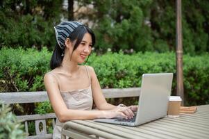 ein attraktiv asiatisch Frau ist Arbeiten auf ihr Laptop Computer beim ein Tabelle im ein Grün Garten. foto