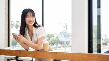 ein attraktiv asiatisch Frau ist halten ihr Telefon, lächelnd Weg von das Kamera, chillen im ein Cafe. foto