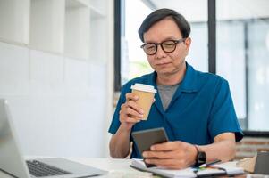 ein erfahren Senior asiatisch männlich Innere Designer ist nippen Kaffee während Arbeiten beim seine Schreibtisch. foto