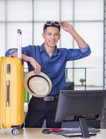 jung Mann Stehen im seine Büro suchen hoch, lächelnd, und berühren groß Gelb Gepäck. bereiten zum das bevorstehende Urlaub. Urlaub Konzept foto