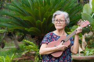 heiter ein Senior Frau spielen das Ukulele während Stehen im ein Garten. entspannend durch Singen und abspielen klein Gitarre glücklich und genießen Leben nach in den Ruhestand gehen. Konzept von alt Menschen und Gesundheitswesen foto