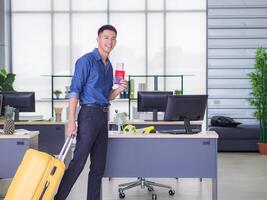 jung Mann Stand im Büro suchen beim das Kamera und lächeln, links Hand zeigen ein Anerkennung Karte und richtig Hand halten groß Gelb Gepäck. bereiten zum das bevorstehende Urlaub. Raum zum Text. Urlaub Konzept foto