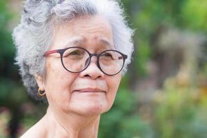 ein Porträt von ein Alten Frau tragen Brille und suchen Weg während Stehen im ein Garten. Raum zum Text. Konzept von alt Menschen und Gesundheitswesen foto