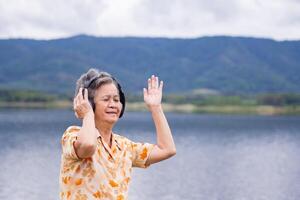 Porträt von Senior asiatisch Frau tragen kabellos Kopfhörer Hören zu ein Liebling Lied während Sitzung auf ein hölzern Stuhl Seite das Teich. Konzept von alt Menschen und Entspannung foto