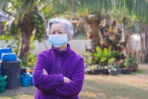 Senior Frau Waffen gekreuzt, tragen ein Gesicht Maske und suchen beim das Kamera während Stehen im ein Garten. Maske zum Schutz COVID-19, Virus, Bakterien, usw. Konzept von alt Menschen und Gesundheitswesen foto