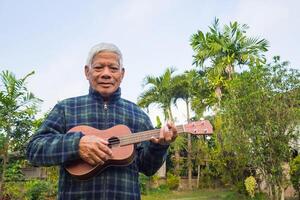 Porträt von ein glücklich Senior Mann spielen das Ukulele, lächelnd und suchen beim das Kamera während Stehen im ein Garten. Raum zum Text. Konzept von alt Menschen und Entspannung foto