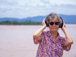 Porträt von ein glücklich Alten Frau tragen kabellos Kopfhörer Hören zu Musik- während Stehen Seite das Fluss. Raum zum Text. Konzept von alt Menschen und Entspannung foto