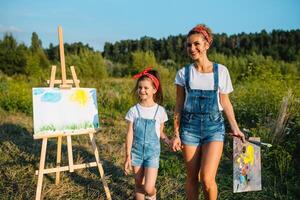 Schönheit Mutter Farbe mit ihr wenig Tochter. stilvoll Frau Zeichnung das Bild mit wenig Mädchen. süß Kind im ein Weiß T-Shirt und Blau Jeans. Mutter Tag. foto