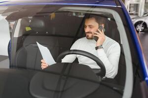 bärtig Geschäftsmann mit Zelle Telefon, bärtig Mann im stilvoll Jacke, Mann im Sonnenbrille, Mann im Luxus Auto, Frisur, hübsch, attraktiv, reich, Hand betrachten, Jeans, beschäftigt, Telefon arbeiten, Designer, glücklich foto