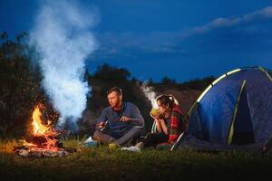 jung Paar haben ein sich ausruhen beim Lagerfeuer neben Lager und Blau Tourist Zelt, Trinken Tee, genießen Nacht Himmel. das Konzept von aktiv Erholung und Reise mit ein Zelt foto