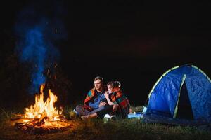 Nacht Camping im das Berge. glücklich Paar Reisende Sitzung zusammen neben Lagerfeuer und glühend Tourist Zelt. auf Hintergrund groß Felsblock, Wald und Nacht Himmel. foto