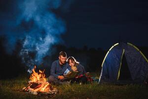 Nacht Camping im das Berge. glücklich Paar Reisende Sitzung zusammen neben Lagerfeuer und glühend Tourist Zelt. auf Hintergrund groß Felsblock, Wald und Nacht Himmel. foto