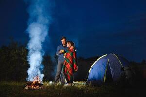 Nacht Camping im das Berge. glücklich Paar Reisende Sitzung zusammen neben Lagerfeuer und glühend Tourist Zelt. auf Hintergrund groß Felsblock, Wald und Nacht Himmel. foto