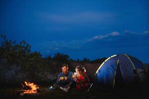 glücklich romantisch Paar Reisende ruhen beim Lagerfeuer in der Nähe von glühend Tourist Zelt unter tolle Nacht Himmel. das Konzept von aktiv Erholung und Reise mit ein Zelt foto