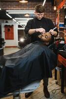 Seite Aussicht von ernst Mann mit stilvoll modern Haarschnitt suchen nach vorne im Barbier Geschäft. Hand von Barbier behalten Gerade Rasierer und Schneiden modisch Streifen auf Kopf von Klient. foto