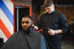 Nahansicht von Prozess von Trimmen von Haar im Barbier Geschäft. qualifiziert Barbier behalten Clipper im Hände und korrigieren gestalten von Haar zu männlich Klient Sitzung auf Stuhl. Konzept von Haarschnitt und Rasieren. foto