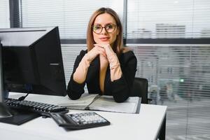 hübsch, Hübsch, Niedlich, perfekt Frau Sitzung beim ihr Schreibtisch auf Leder Stuhl im Arbeit Bahnhof, tragen Gläser, formelle Kleidung, haben Laptop und Notizbuch auf das Tabelle foto