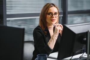 hübsch, Hübsch, Niedlich, perfekt Frau Sitzung beim ihr Schreibtisch auf Leder Stuhl im Arbeit Bahnhof, tragen Gläser, formelle Kleidung, haben Laptop und Notizbuch auf das Tabelle foto